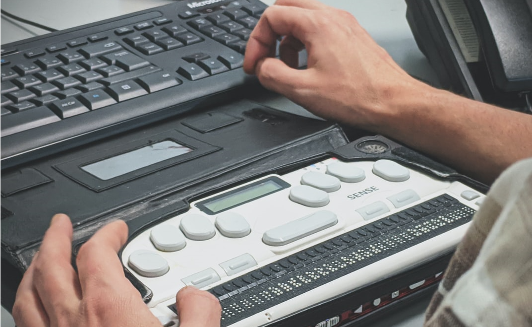 persons hands on a desk with a keyboard and a piece of accessibility equipment, a Braille reader.